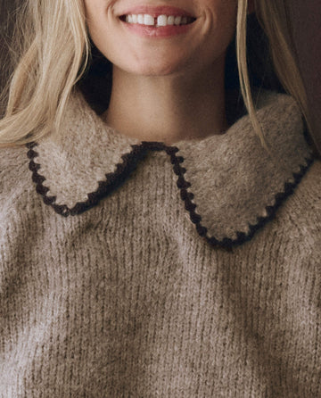 A woman with blonde hair wears THE GREAT. F25 D1 The Crochet Collar Pullover in Oat with Brown, a beige knit sweater featuring a relaxed fit and large brown-edged wool collar, smiling slightly.
