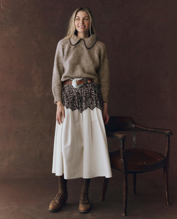 A woman stands indoors, smiling near a wooden chair in front of a brown backdrop, wearing THE GREAT. F25 D1's The Regency Skirt in Cream With Western Lace Print, a tan sweater, patterned scarf belt, and brown lace-up boots.