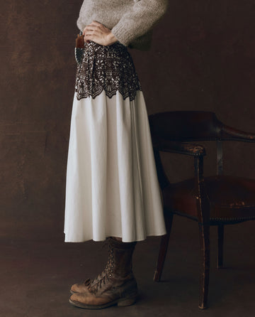 A person stands beside a wooden chair, wearing a beige sweater, brown lace-up boots, and THE GREAT. F25 D1's The Regency Skirt in cream with Western lace print, hands resting on their hips.