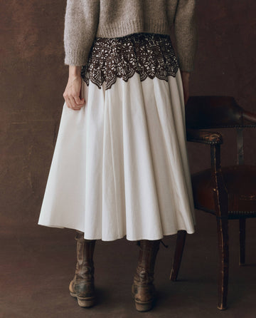 A person stands next to a brown leather chair wearing a beige sweater, brown lace-up boots, and THE GREAT. F25 D1's The Regency Skirt in cream with Western lace print.