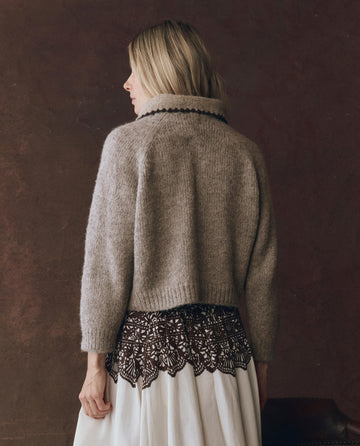 A blonde person wears THE GREAT. F25 D1 "The Crochet Collar Pullover" in oat with brown, styled with a white skirt with a brown lace overlay, standing against a dark brown background.