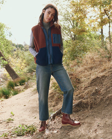 Standing outdoors on a dirt path, a person wears jeans, a gray long-sleeve shirt, brown boots, and THE GREAT. F25 TGO's The Plush Tundra Vest in Chocolate & Navy. Trees and foliage appear in the background.