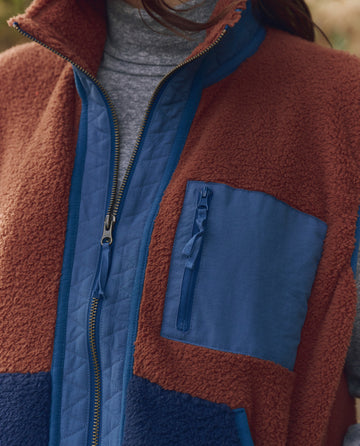 Close-up of a person wearing THE GREAT. F25 TGO's Plush Tundra Vest in Chocolate & Navy, featuring a zipper front and blue chest pocket over a gray turtleneck—evoking vintage camping style.