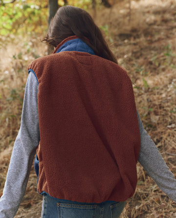 A person with long hair, seen from behind, walks outdoors in dry grass and trees while wearing THE GREAT. F25 TGO The Plush Tundra Vest in Chocolate & Navy and blue jeans.