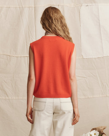 A person with wavy, light brown hair is seen from behind in THE GREAT. PS26 The Cashmere Pullover Vest in Hot Orange, paired with white jeans, standing before a beige fabric backdrop.