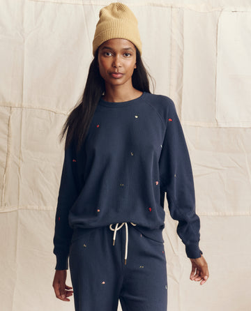 A woman wearing THE GREAT. PS26's The College Sweatshirt, embroidered in navy with tossed floral details, stands before a beige fabric backdrop in Los Angeles.