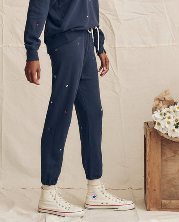 Wearing THE GREAT. PS26 The Stadium Sweatpant in navy with tossed floral embroidery, paired with a matching top and white Converse, this cozy LA-made look stands beside a wooden crate adorned with white flowers.