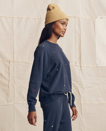 Woman in a tan beanie and THE GREAT. PS26 The College Sweatshirt. Embroidered—Navy with Tossed Floral Embroidery, standing in profile before a cream fabric backdrop, channeling effortless Los Angeles style.