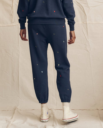 Wearing THE GREAT. PS26 The Stadium Sweatpant in navy with tossed floral embroidery, styled with a matching sweatshirt and white high-top sneakers, against a beige backdrop—effortless LA-made vintage style.