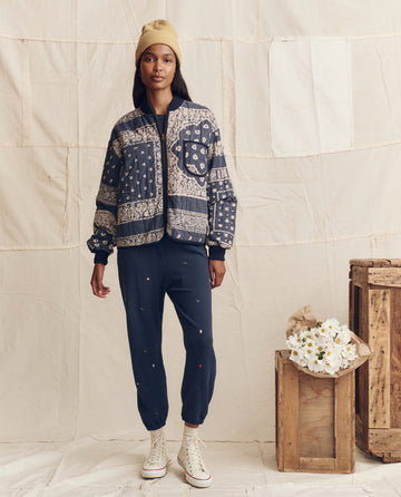 Indoors, a person wears THE GREAT. PS26's The Stadium Sweatpant. Embroidered in navy with tossed floral embroidery, paired with a patterned jacket, white high-top sneakers, and a beige beanie; wooden crates and white flowers nearby.