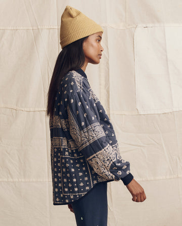 A person stands in profile against a beige fabric backdrop, showcasing THE GREAT. PS26 The Quilted Bomber in Navy Bandana Print and a beige knit beanie for lightweight warmth.