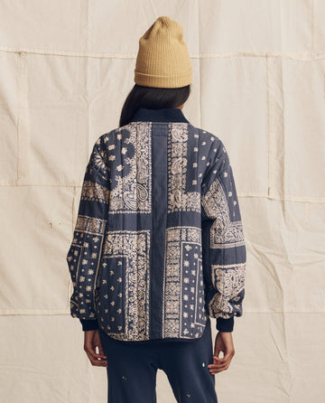 A person in THE GREAT. PS26 The Quilted Bomber -- Navy Bandana Print faces away against a beige fabric backdrop, showcasing the brand's blend of style and lightweight warmth.