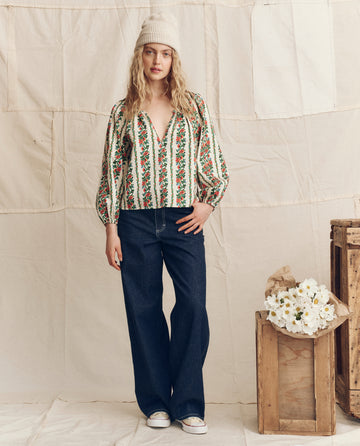 A woman poses indoors beside wooden crates of white flowers, wearing THE GREAT. PS26 The Chariot Top in Tapestry Floral, paired with wide-leg jeans, white sneakers, and a knit hat against a neutral fabric backdrop.