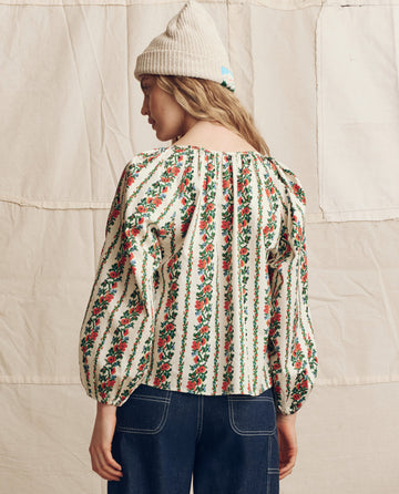 A person stands facing away against a light fabric backdrop, wearing THE GREAT. PS26’s The Chariot Top in Tapestry Floral—a puffed-sleeve blouse—paired with a cream knit beanie and dark blue jeans.