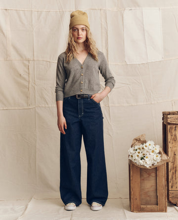 A woman wears THE GREAT. PS26 The Cashmere Tiny V Neck Cardigan in Heather Grey, styled with wide-leg jeans, white sneakers, and a yellow beanie, standing indoors by a wooden crate with white flowers against a beige backdrop.