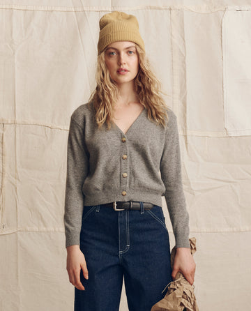 A person in blue jeans, a beige knit hat, and THE GREAT. PS26's Cashmere Tiny V Neck Cardigan in Heather Grey stands before a neutral fabric backdrop, holding a crumpled brown paper bag.