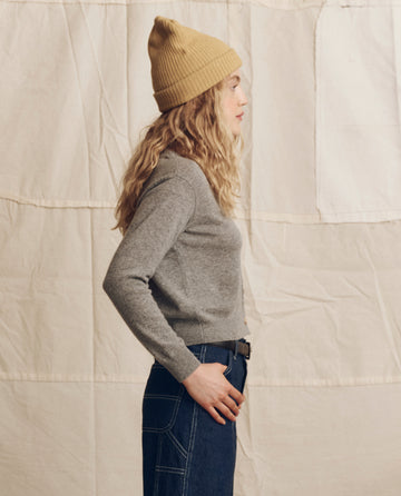 A person with long, wavy blonde hair in a yellow beanie and blue jeans models THE GREAT. PS26 The Cashmere Tiny V Neck Cardigan in Heather Grey, standing in profile before a beige fabric backdrop.