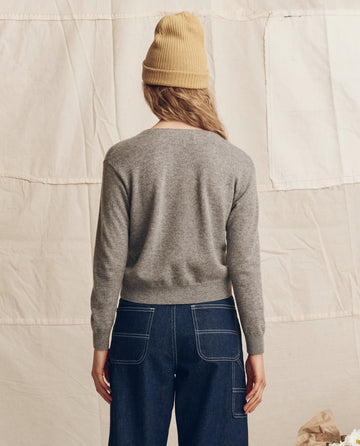A person stands with back to camera in a tan beanie, blue jeans with white stitching, and THE GREAT. PS26 The Cashmere Tiny V Neck Cardigan in Heather Grey, posed against a beige fabric backdrop.