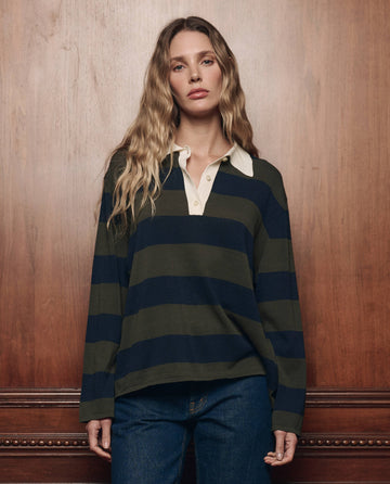 A woman with long wavy hair, wearing THE GREAT. F26 COLLECTORS’ The Academy Polo Sweater in Army League Stripe—a dark green and navy striped rugby sweater with a white collar—paired with jeans, stands in front of a wooden wall.