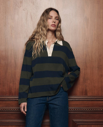 A woman with long, wavy hair stands against a wooden wall, wearing THE GREAT. F26 COLLECTORS The Academy Polo Sweater in Army League Stripe and blue jeans.