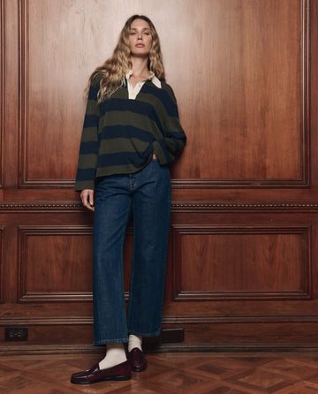 A person stands against a wood paneled wall, wearing the Academy Polo Sweater in Army League Stripe from THE GREAT. F26 COLLECTORS layered under a dark striped rugby shirt, paired with wide-leg jeans, white socks, and loafers.