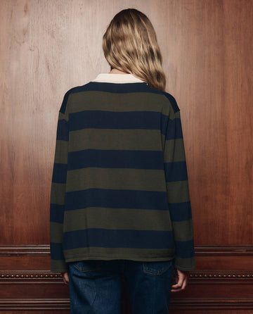 A person with long, wavy hair stands facing a wooden wall, wearing THE GREAT. F26 COLLECTORS The Academy Polo Sweater -- Army League Stripe and blue jeans.