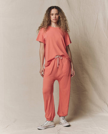 A woman stands against a neutral backdrop wearing THE GREAT. SU26 SEASONAL CORE The Jersey Jogger Pant in Guava, paired with a loose coral short-sleeve top and white sneakers.