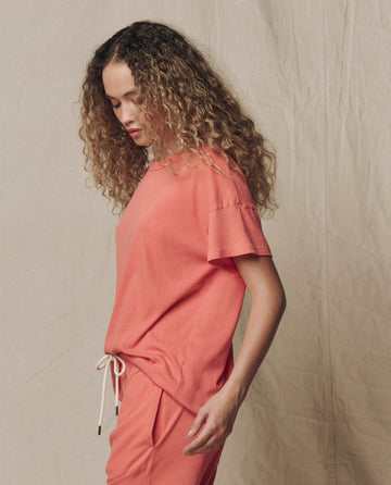 A person with curly hair wears THE GREAT. SU26 SEASONAL CORE The Boxy Crew. Solid -- Guava, standing in profile against a neutral fabric backdrop.