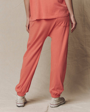 A person faces away from the camera, wearing THE GREAT.'s The Jersey Jogger Pant in Guava (SU26 SEASONAL CORE) and white sneakers, standing against a neutral fabric backdrop.