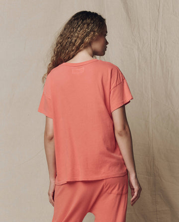 Person with long curly hair, seen from behind, wears THE GREAT. SU26 SEASONAL CORE The Boxy Crew. Solid in Guava with matching pants, standing against a neutral fabric backdrop.