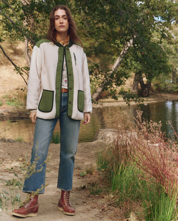 A woman stands by a pond surrounded by trees, wearing THE GREAT. F25 TGO’s The Plush Tundra Jacket in Cream & Army, paired with blue jeans and brown boots.