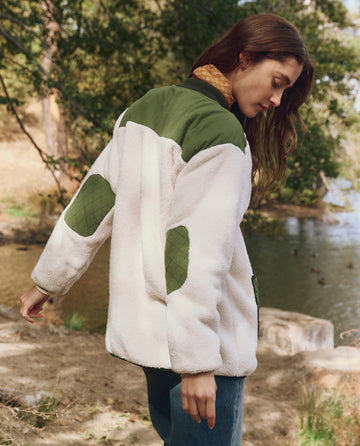 A woman wears The Plush Tundra Jacket in Cream & Army by THE GREAT. F25 TGO as she stands outdoors near a tranquil lake, surrounded by trees and nature.