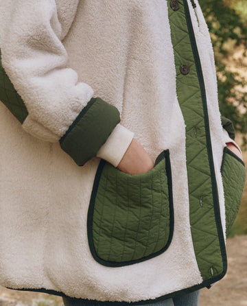 Wearing THE GREAT. F25 TGO's Plush Tundra Jacket in Cream & Army—plush fleece with green quilted trim and multiple pockets—outdoors with hands tucked in, ideal for vintage camping gear fans.