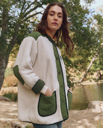 A person wears THE GREAT. F25 TGO The Plush Tundra Jacket in Cream & Army, featuring multiple pockets, standing outdoors near trees and a pond with hands in pockets, looking at the camera.