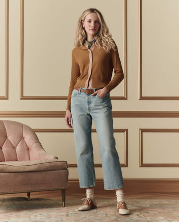 A woman stands indoors beside a pink sofa, wearing THE GREAT. SP26 D2 The Dainty Cardigan in Suntan, light blue jeans, white socks, brown shoes, and a scarf against a beige and gold-paneled wall.
