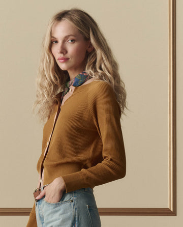 A woman with long, wavy blonde hair wears THE GREAT. SP26 D2's The Dainty Cardigan in Suntan over a colorful scarf and blue jeans, standing against a beige background with a picture frame.