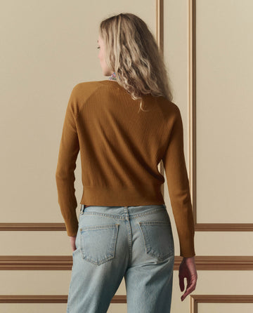 A person with wavy blonde hair, wearing THE GREAT. SP26 D2 The Dainty Cardigan in Suntan and light blue jeans, stands with their back to the camera against a beige wall with molding.