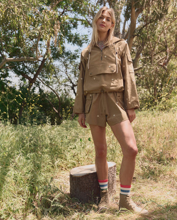 A woman stands outdoors near a tree stump, wearing THE GREAT. SP26 TGO's The Embroidered Cargo Short in Khaki With Cream Daisies, paired with a tan jacket, striped socks, and tan boots amid trees and greenery.
