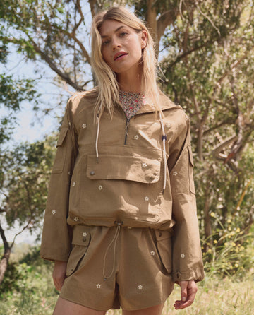 A woman stands outdoors in THE GREAT. SP26 TGO Embroidered Cargo Short—khaki with cream daisies—paired with a tan long-sleeve jacket featuring floral embroidery. Trees and sunlight fill the background.