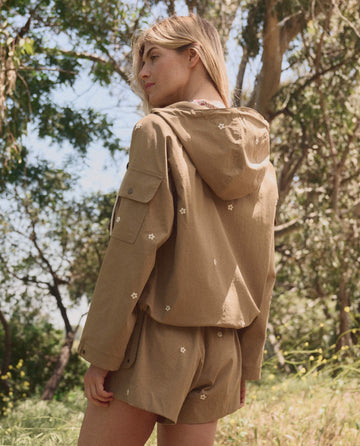 Outdoors among trees, a person wears THE GREAT. SP26 TGO's The Embroidered Cargo Short in khaki with cream daisies, paired with a light brown hooded jacket.