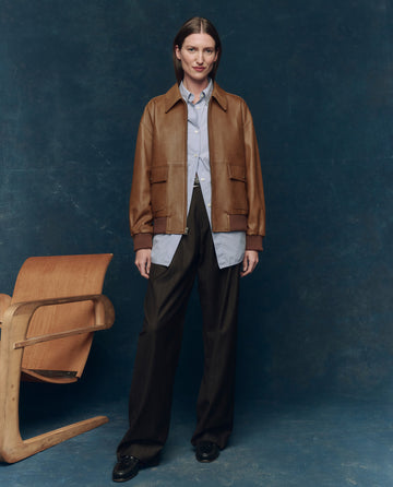 Indoors against a dark blue backdrop, a person wears THE GREAT. F25 COLLECTORS The Flight Jacket in Saddle Brown over a blue button-up shirt, dark wide-leg trousers, and black shoes, standing by a wooden chair.