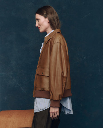 A person stands in profile against a dark blue background, wearing THE GREAT. F25 COLLECTORS The Flight Jacket in Saddle Brown over a blue button-up shirt and dark pants.