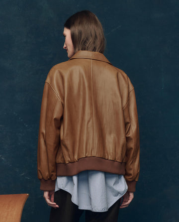 A person with shoulder-length brown hair, back to the camera, wears THE GREAT. F25 COLLECTORS’ The Flight Jacket in Saddle Brown over a blue striped shirt.