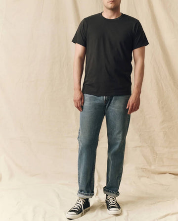 A person stands against a beige fabric backdrop, wearing THE GREAT. FALL 23 MEN The Men's Pure Knits Slim Tee in Solid Almost Black, paired with blue jeans and black high-top sneakers.