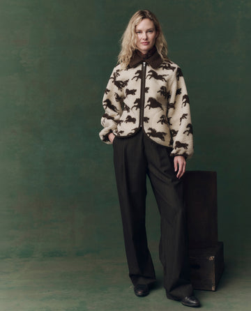 A person stands against a green background, wearing THE GREAT. F25 D2’s The Pasture Jacket in Cream with Hickory, paired with dark wide-leg trousers and black shoes.