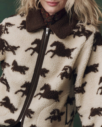 A person in THE GREAT. F25 D2’s The Pasture Jacket in Cream with Hickory, featuring a plush zip-up design and horse pattern, showcases warmth and style with its cozy brown collar.