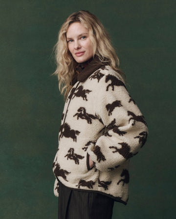 A person with wavy blond hair models THE GREAT. F25 D2's The Pasture Jacket in Cream with Hickory, standing against a plain dark green background.