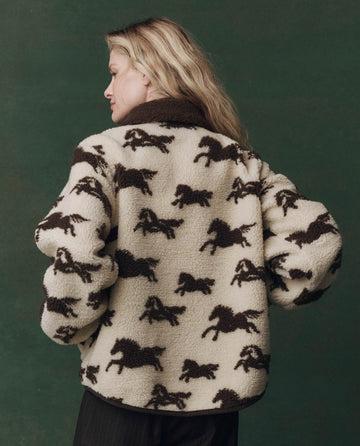 A person wears THE GREAT.'s The Pasture Jacket in Cream with Hickory, featuring a horse pattern, standing against a dark green background and facing away from the camera.