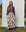 A person stands outdoors in front of a light blue backdrop, casually dressed in THE GREAT. PS25's The Shrunken Henley in Capital Stripe and a floral print long skirt.