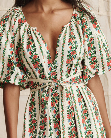 A woman wears THE GREAT. PS26 The Fairground Dress—Tapestry Floral, a cream pullover with red and green floral patterns, puffed sleeves, deep V-neckline, and a matching fabric belt at the waist.